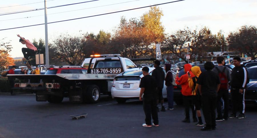 A teenager ollies off the back of a tow truck parked in the plaza while spectators watch and record the stunt. 