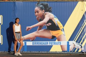 Varsity player Cassia Ramelb smiles next to the new banner across the track field which features her jumping across a hurdle. The junior has been hurdling for Birmingham Community Charter High School track and field team for three years and plans to continue to through college.
